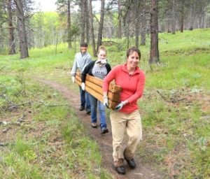 Classroom Without Walls instructor Alli Neils-Lemone (in the lead) helps shoulder the load as she and students Mollie DuBose and Teran Koerner carry in some timber beams that were used in the replacement of a pedestrian footbridge over the creek that had been damaged by falling timber. Michael Howell photo.